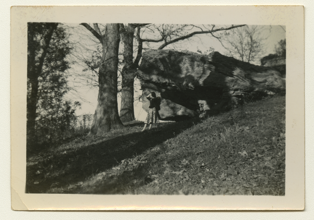 A black and white long-shot photograph of Anne Martin Wilson posing under a large rock in a grassy area