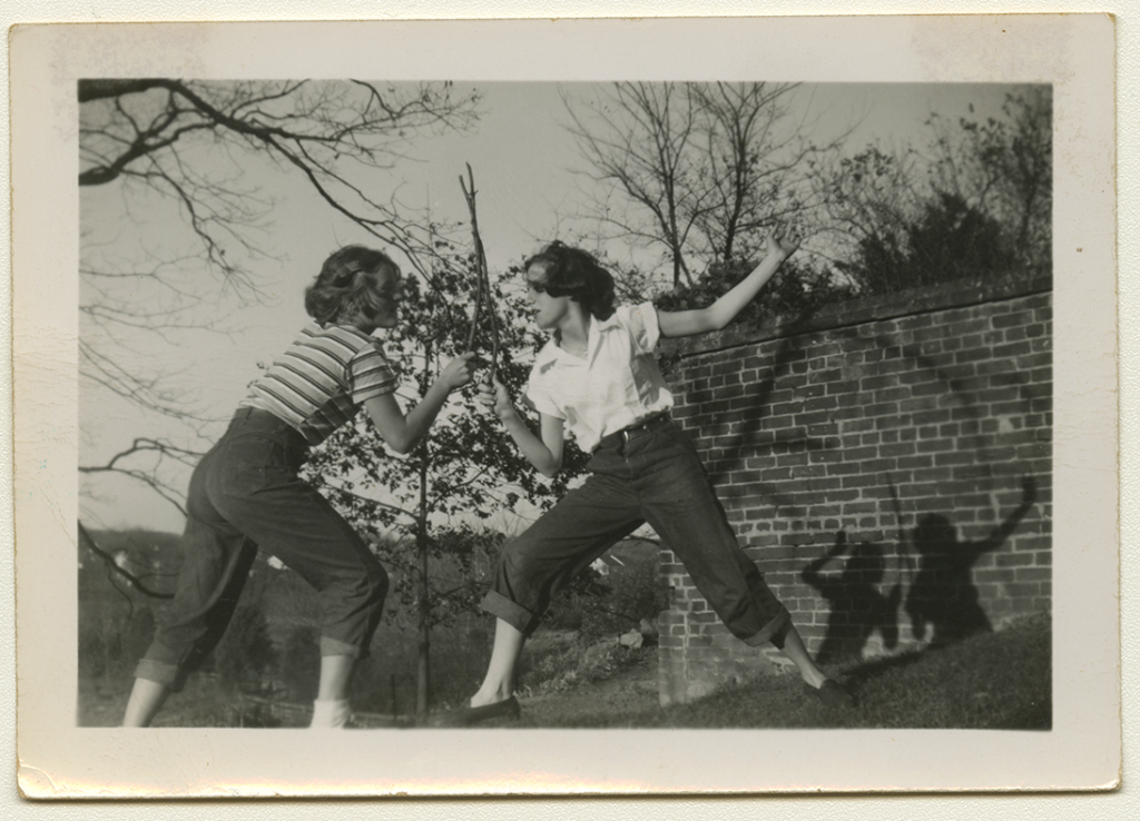 A black and white photo of a young Alma and Anne Rowe "sword fighting" with sticks next to a brick wall